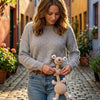 A woman in a sweater holds a handmade crochet sloth and squirrel plush charm outdoors on a cobblestone street.