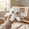 A hand is holding a white flip octopus plush toy against a blurred background of a window and wooden crib.