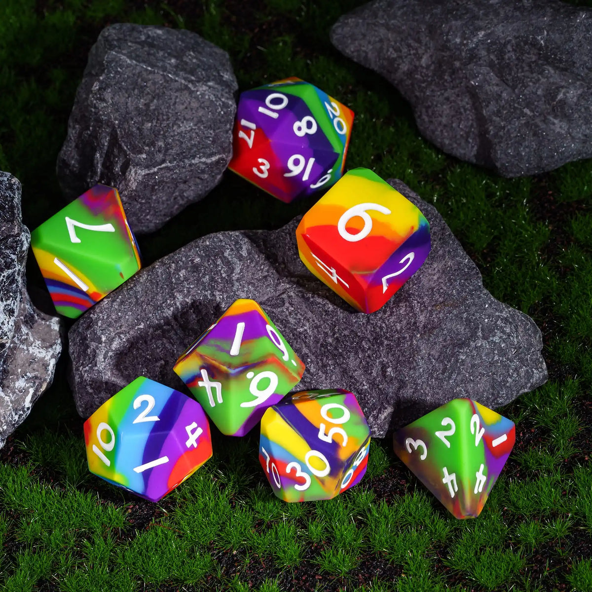A set of colorful silicone polyhedral dice resting on green grass among dark rocks.