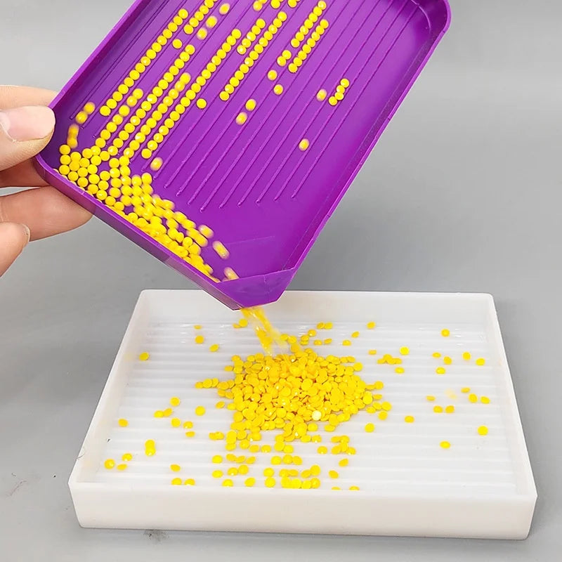 A hand places yellow beads onto a white tray, demonstrating the use of a purple diamond painting tool kit.