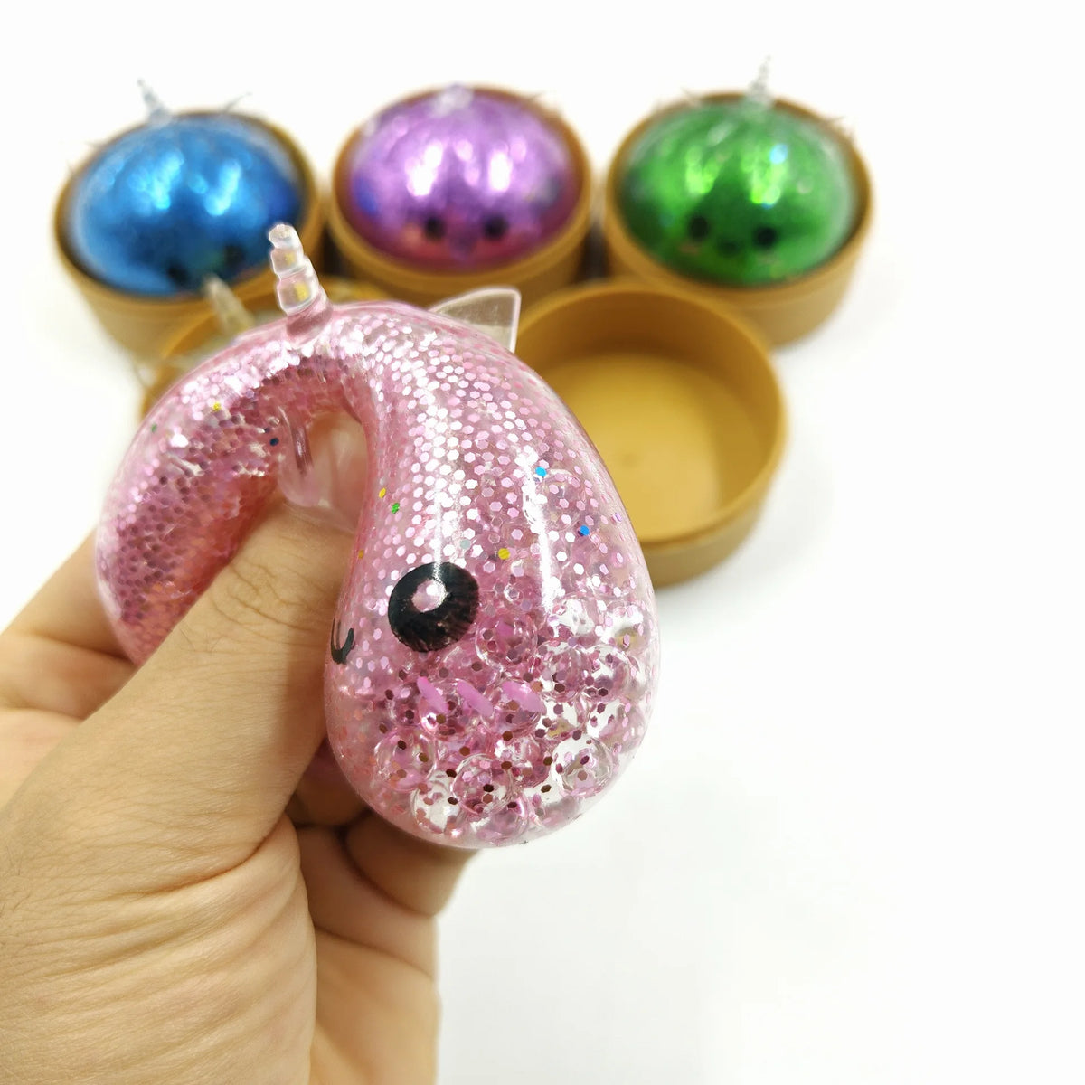 A hand holds a pink glitter unicorn dumpling squishy with a small steamer box, surrounded by other colorful squishies.