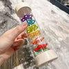 A hand holds a wooden rain sound shaker toy with colorful plastic raindrops inside.