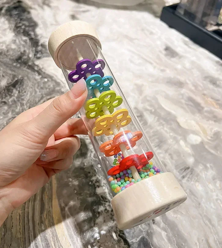 A hand holds a wooden rain sound shaker toy with colorful plastic raindrops inside.