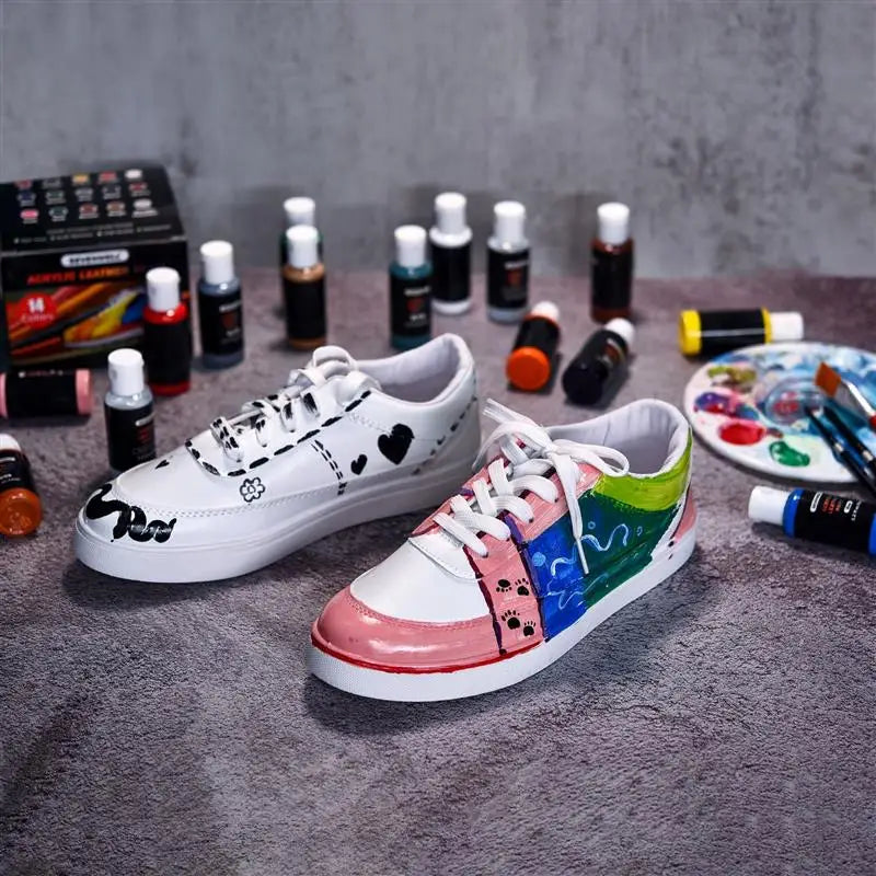 A pair of white and pink acrylic paint-splattered sneakers displayed on a gray fabric surface with various paint bottles.