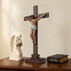 A wooden crucifix made of building blocks with a figure of Jesus and an angel beside it on a dark brown wooden surface.