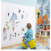A young child sits on a yellow stool in front of a white wall decorated with chalk drawings and stickers.