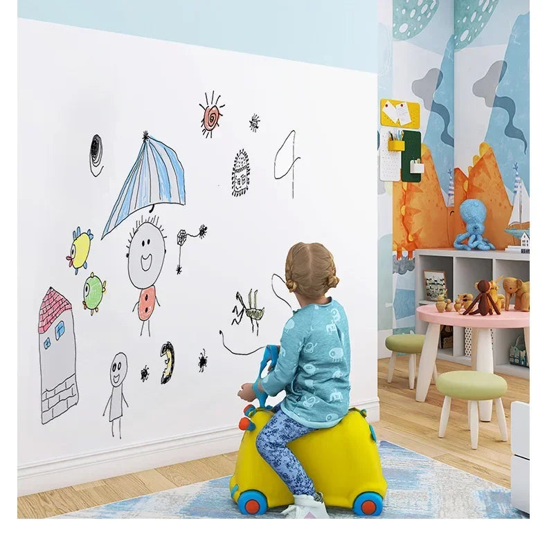 A young child sits on a yellow stool in front of a white wall decorated with chalk drawings and stickers.