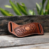 A handcrafted brown leather knife sheath with a decorative pattern and a belt loop resting on weathered wood.