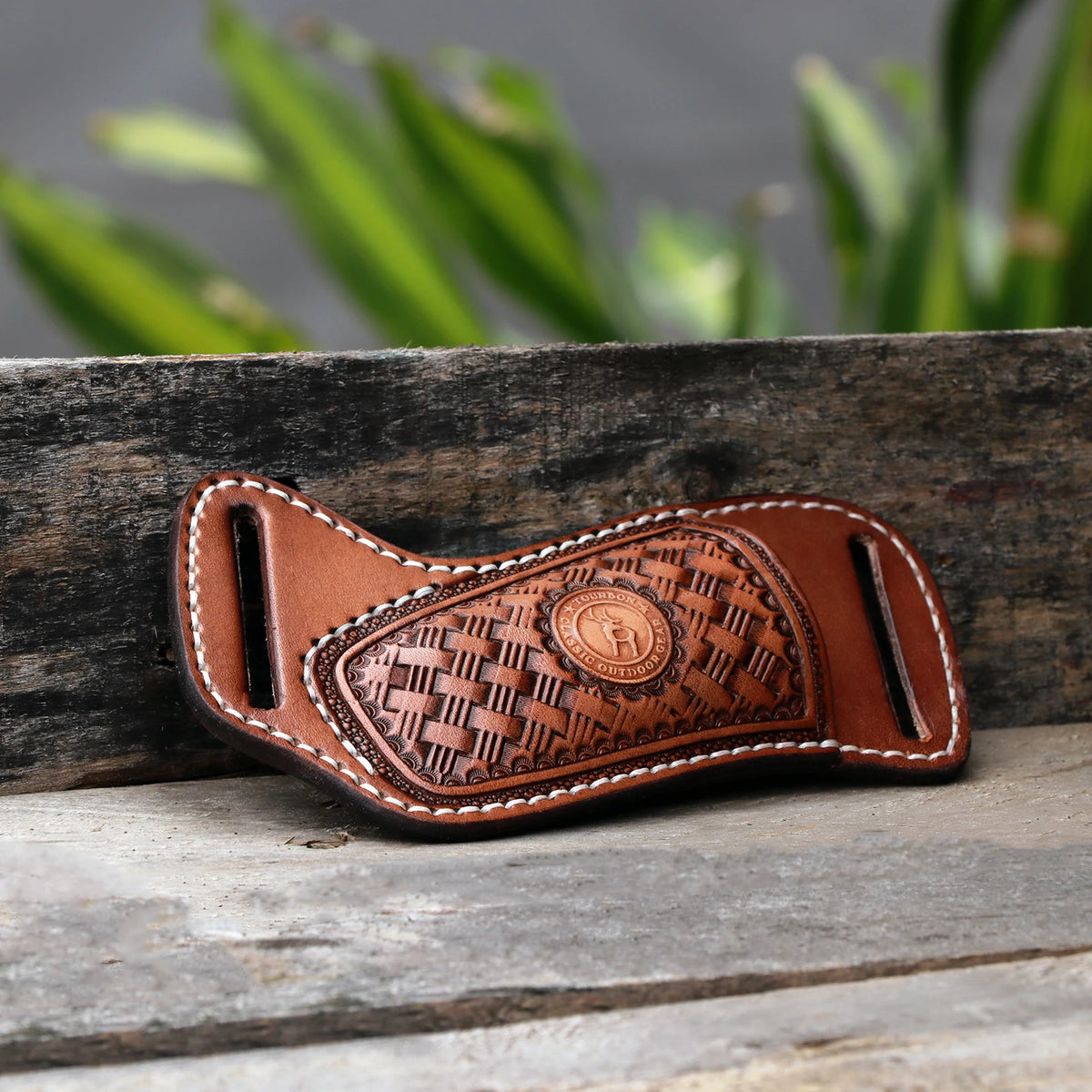 A handcrafted brown leather knife sheath with a decorative pattern and a belt loop resting on weathered wood.