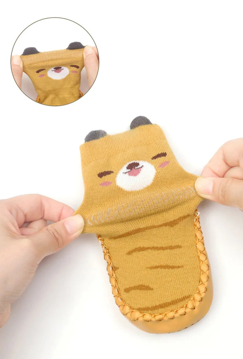 A hand holds a cute cartoon bear non-slip sock for babies and toddlers, featuring brown fur and pink nose.