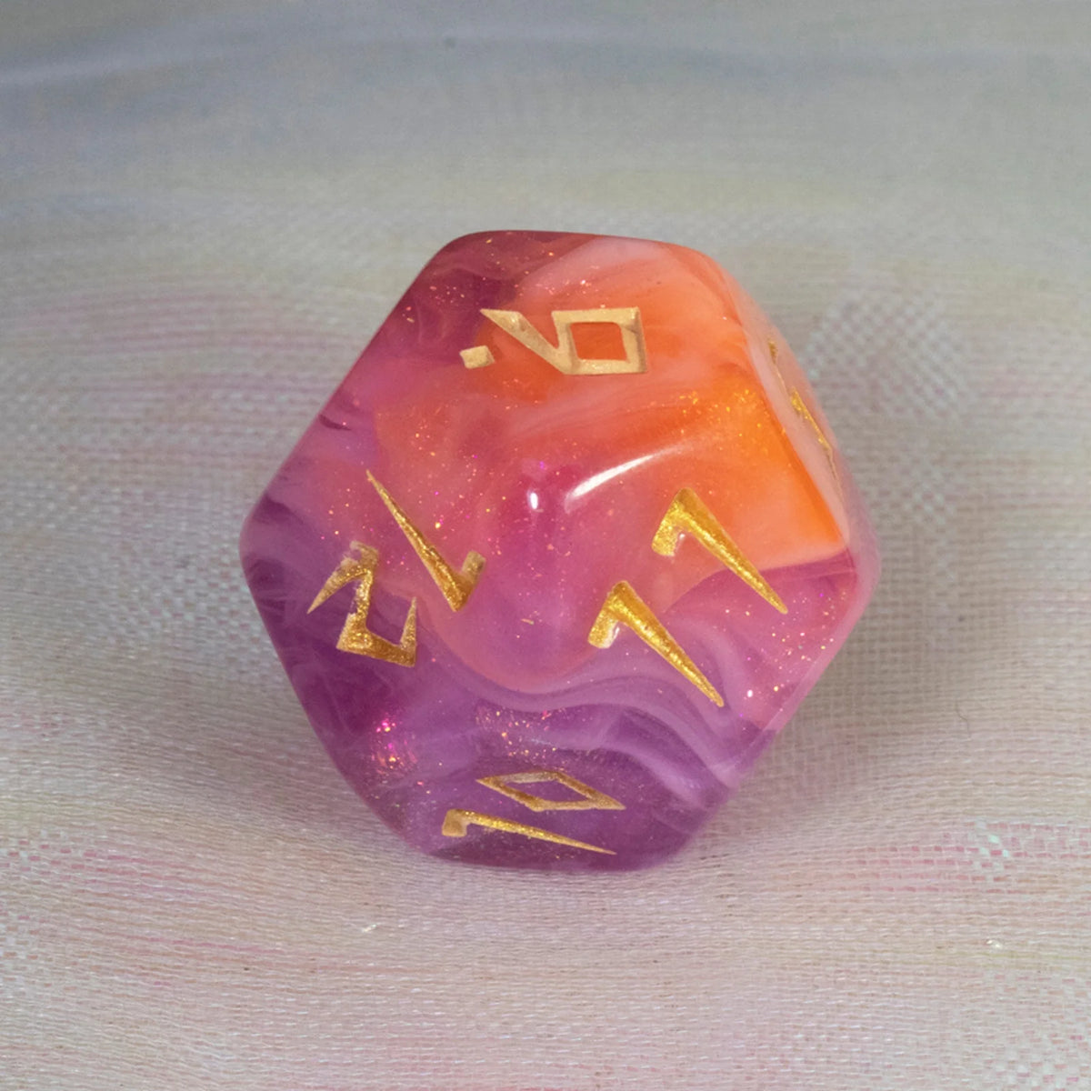 A translucent, faceted polyhedral die with a pink and orange gradient and gold numbers is displayed on a light fabric.