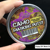 A hand holds a tin of Camo Masking Putty for model painting with a purple and green camouflage pattern visible on the label.
