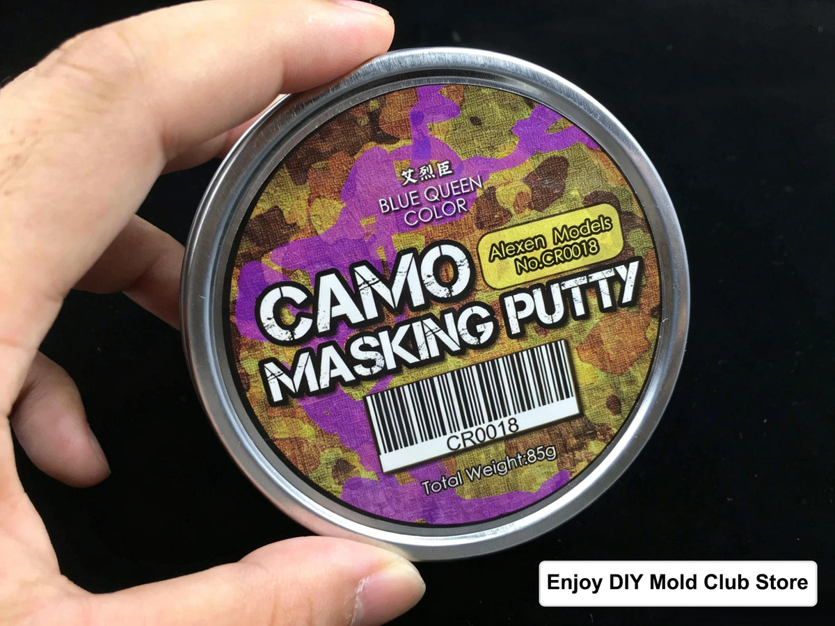 A hand holds a tin of Camo Masking Putty for model painting with a purple and green camouflage pattern visible on the label.