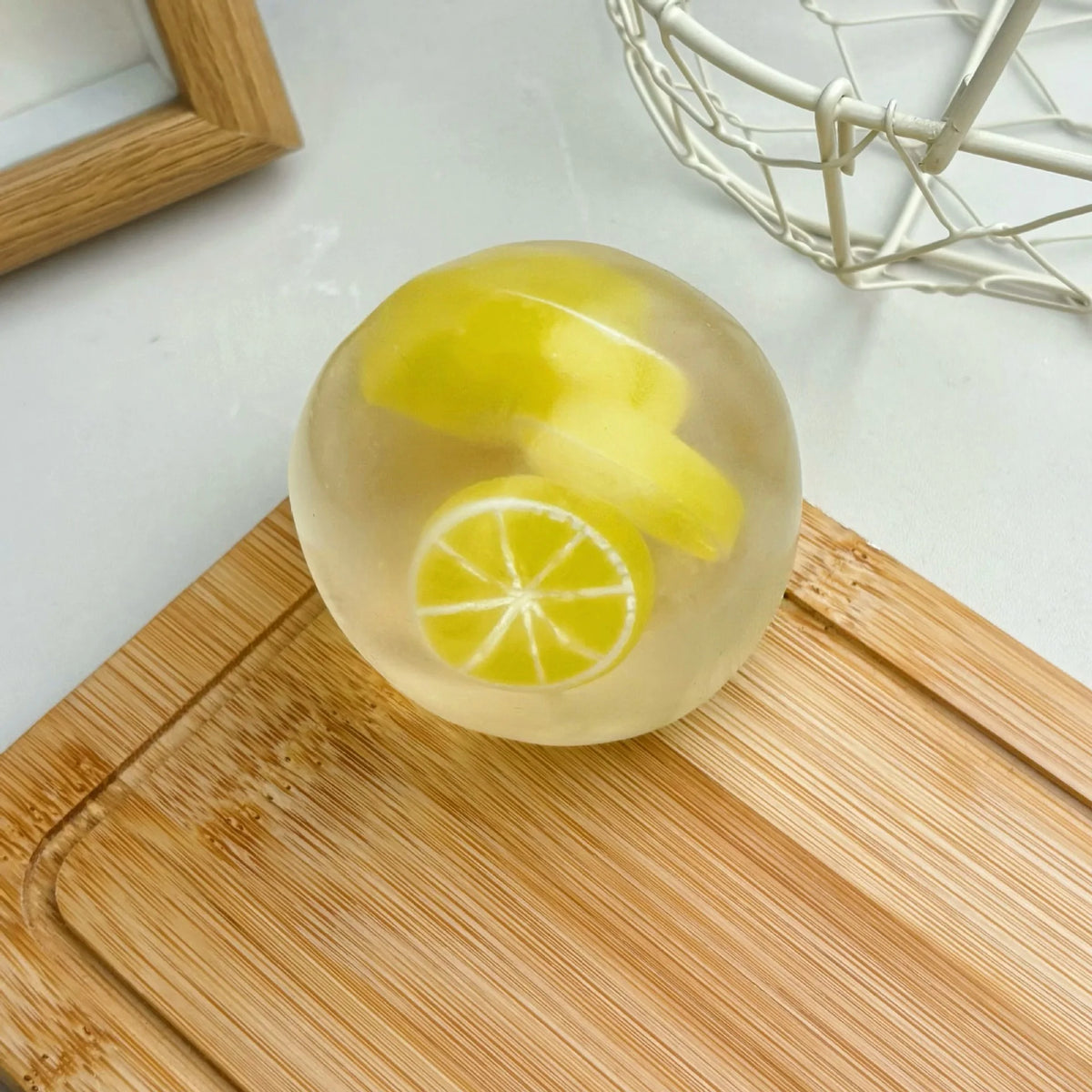 A clear, round squishy stress ball filled with yellow liquid and a slice of lemon, resting on a wooden cutting board.