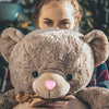 A young girl is holding a fluffy brown teddy bear with a pink nose and safety eyes.