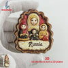 A hand holds a wooden magnet featuring a Russian-inspired scene with multiple faces and the word Russia.