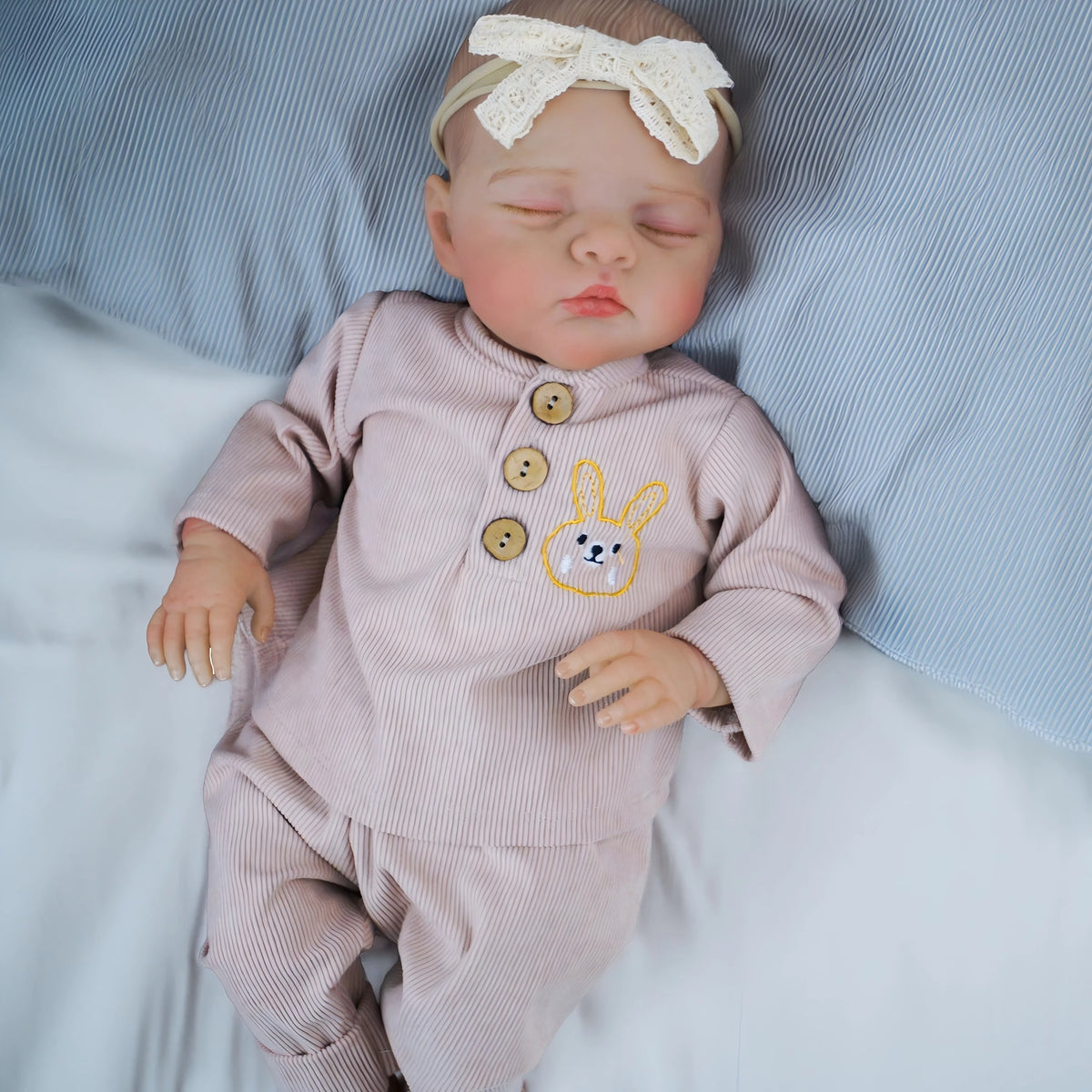 A newborn reborn baby doll wearing a pink outfit and a white bow, sleeping on a blue striped pillow.