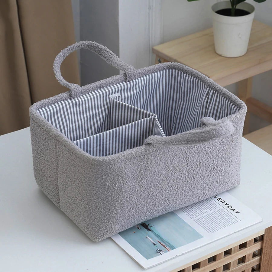 Sac multifonction gris tricoté pour bébé avec poignée, sur une surface blanche à côté d'un magazine.