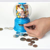 A hand is dispensing silver coins from a blue gumball dispenser filled with colorful candies.