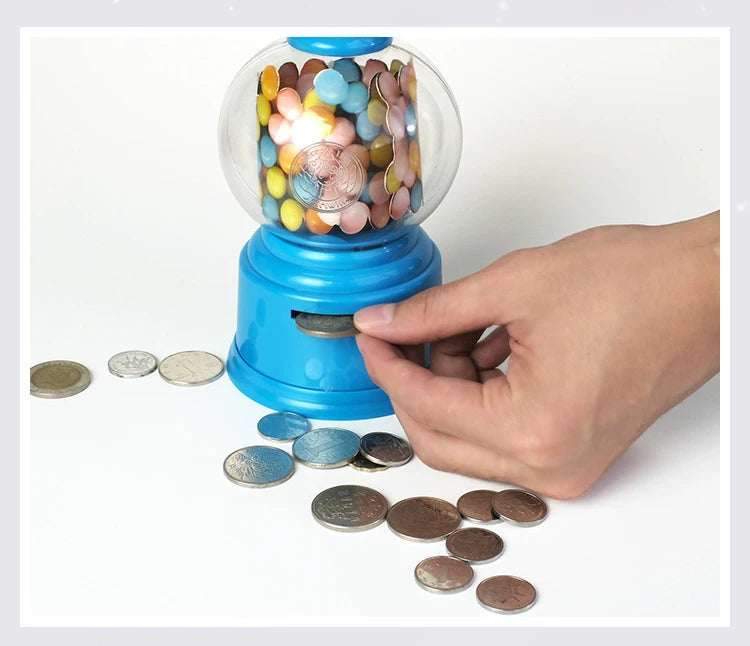 A hand is dispensing silver coins from a blue gumball dispenser filled with colorful candies.