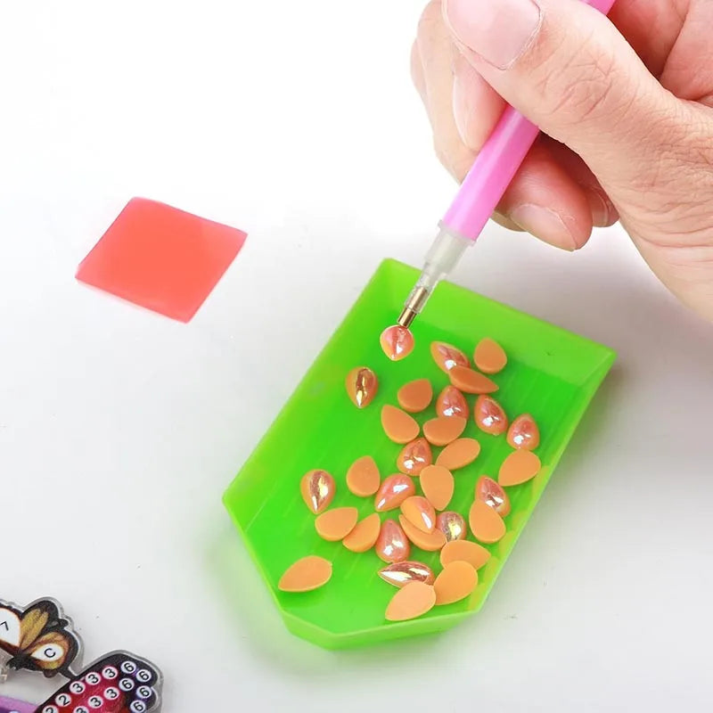 A hand uses a pink drill pen to apply adhesive to small, orange-colored sequins in a green tray for diamond painting.