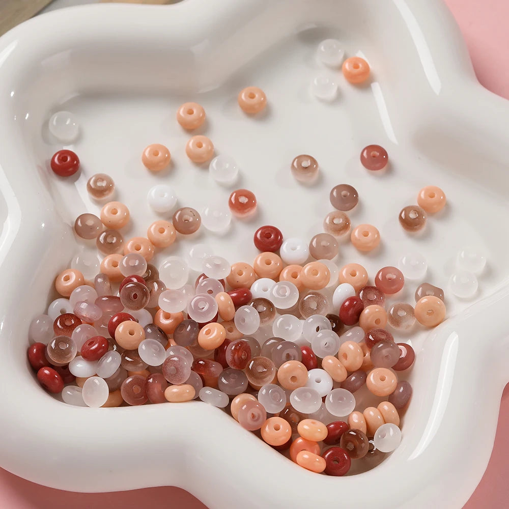 A collection of various colored glass spacer beads, including jade tones and clear beads, are displayed in a white dish.