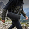 A person wearing a dark jacket is carrying an olive green backpack and a black portable Bluetooth speaker while hiking in a m