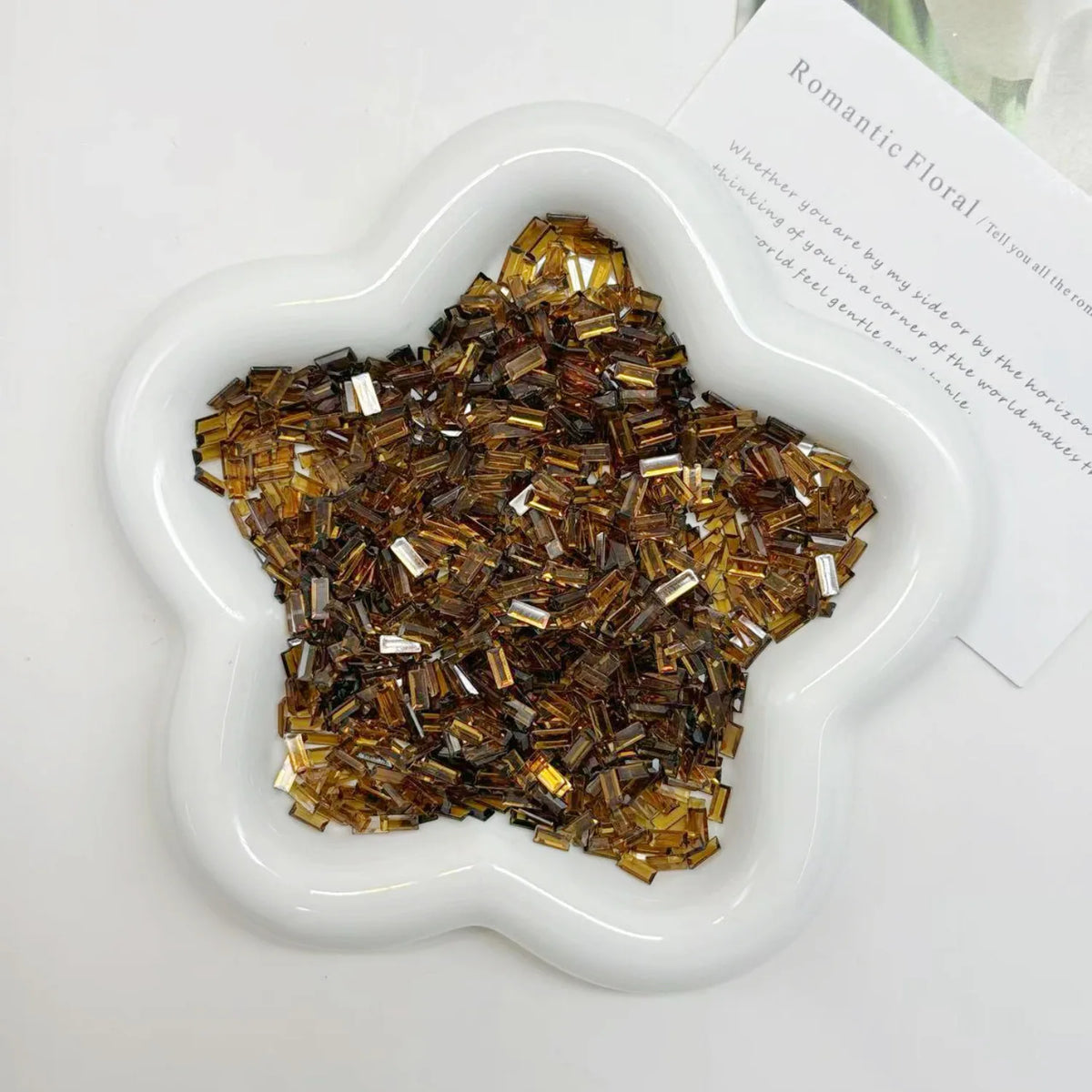 A white dish filled with gold and brown acrylic ice strips for nail art and DIY crafts.