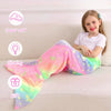 A young girl is sitting on a sofa wearing a bright, glow-in-the-dark mermaid tail blanket for kids.
