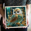A hand holds a framed piece of art featuring a detailed painting of a golden-eyed owl with iridescent blue and green feathers