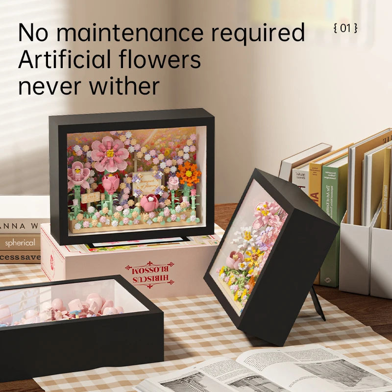 A display of artificial flowers in a black frame, alongside an open box of building blocks and a separate framed flower arran