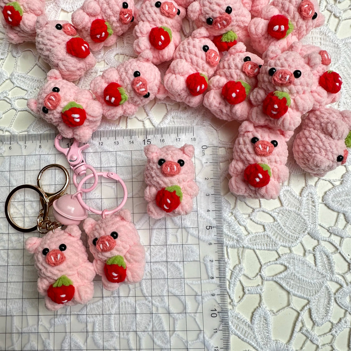 Pink crochet mini pig keychains arranged on a white lace background with a ruler.