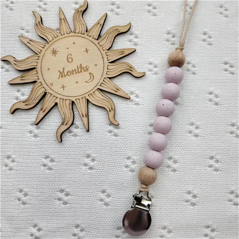 A wooden sun-shaped bead pacifier clip with a string of light purple beads and a silicone teether.