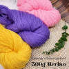 Three skeins of pink, purple, and yellow Merino wool yarn are displayed on a woven mat.
