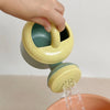 A hand is holding a yellow and teal kids sand and water play set with a pull-along bulldozer trolley over a terracotta bowl.