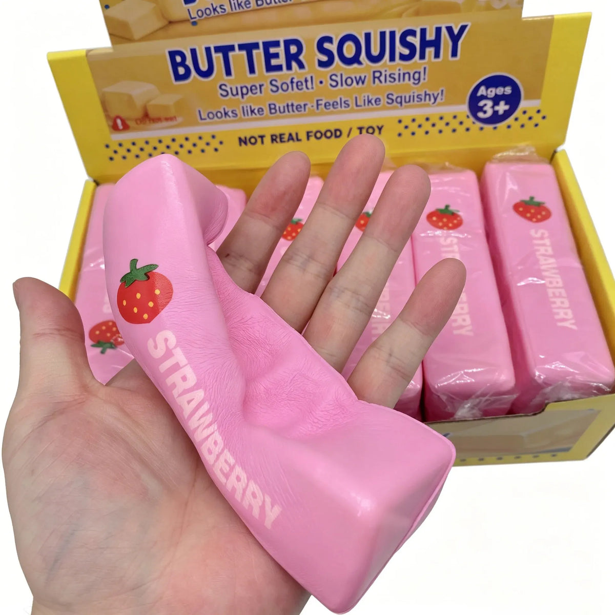 A hand holds a pink strawberry squishy toy inside its original packaging box.