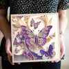 A hand holds a DIY diamond painting kit featuring sparkling purple and silver butterfly art on a clear background.