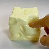 A hand is squeezing a pale yellow, squishy cheese cube stress ball.