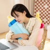 A young woman is holding a light blue milk bottle plush pillow with yellow accents, resting it on a white table.