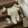 A cream knit ruffle romper for a baby girl displayed on wooden stairs with a matching bow and shoes.