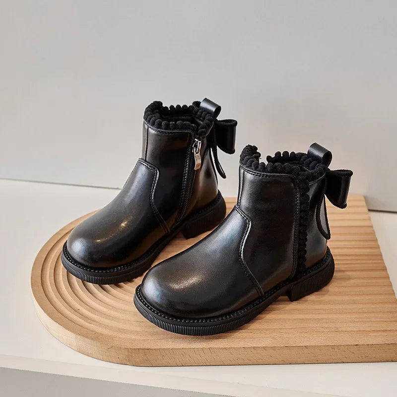 A pair of black ankle boots with a bow-knot detail and side zipper resting on a wooden surface.