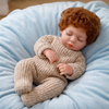 A realistic reborn doll with curly reddish-brown hair wearing a beige knitted outfit lying on a light blue fabric.
