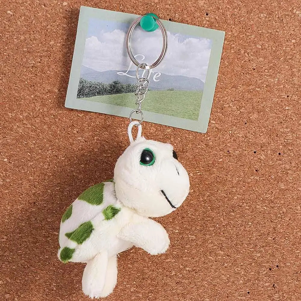 A white and green turtle plush charm attached to a silver keyring, set against a background featuring a landscape.
