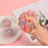 A hand holds a cluster of colorful, translucent squishy stress relief balls against a pink background.