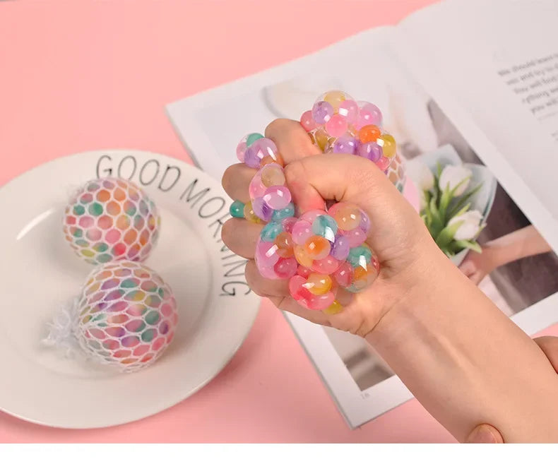 A hand holds a cluster of colorful, translucent squishy stress relief balls against a pink background.