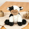 A super soft sitting cow plush toy with black and white markings resting on a beige woven fabric.