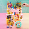 A yellow mini vending machine toy with various snack dispensers and surprise balls displayed on a pink surface.