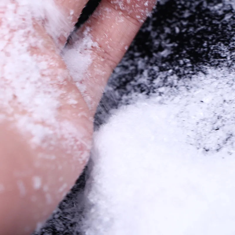 A hand is sprinkling white artificial snow powder onto a dark surface.