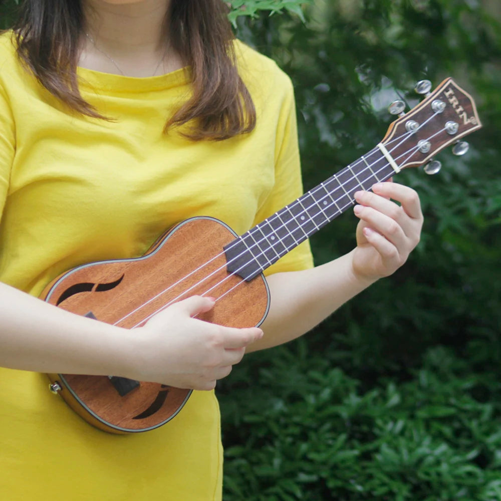 A person holds a brown ukulele with a wooden body and a black neck, set against a green outdoor background.
