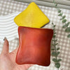 A hand holds two squishy stress relief toys, one yellow and one reddish-orange, on a checkered surface.